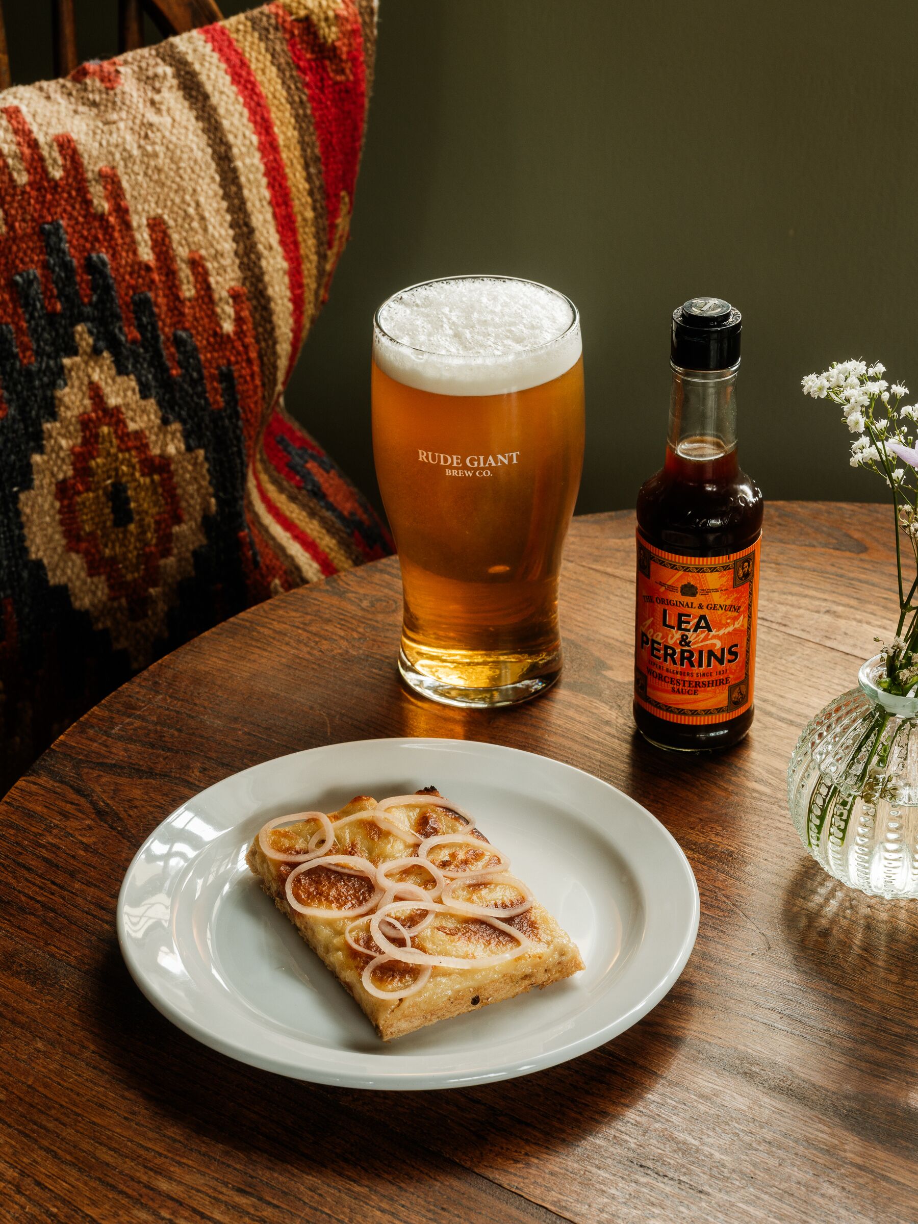 Ein Tisch mit einem Bierglas, einer Flasche Lea & Perrins Sauce und einer Scheibe Zwiebelkuchen auf einem Teller.