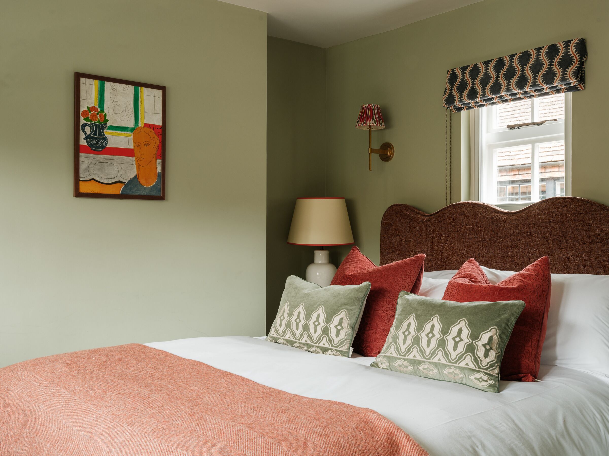 Een slaapkamer met een bed, kussens, een lamp, een schilderij en een raam met een gordijn.