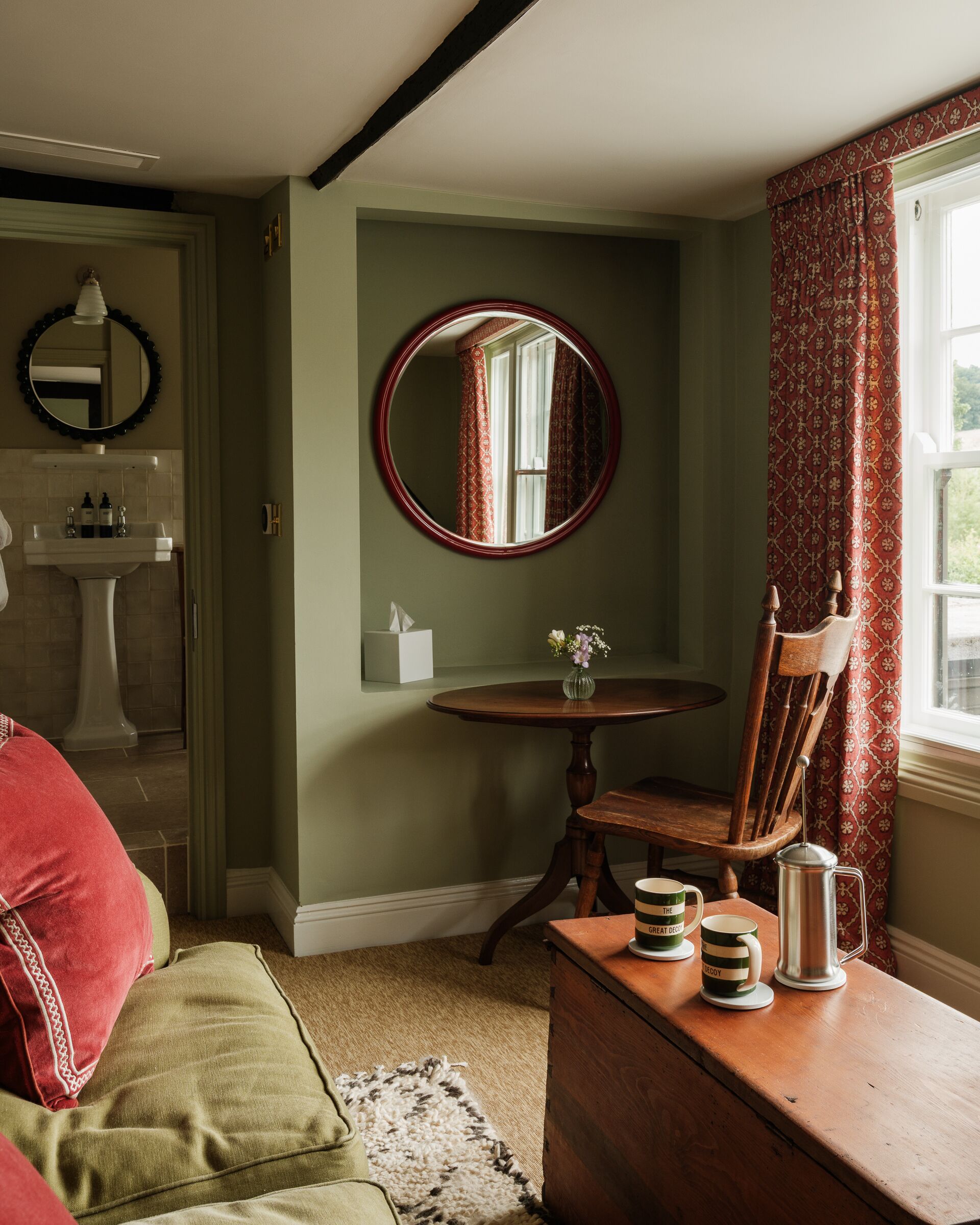 Una habitación con un espejo redondo, una silla de madera, una mesita con un jarrón y una ventana con cortinas estampadas.