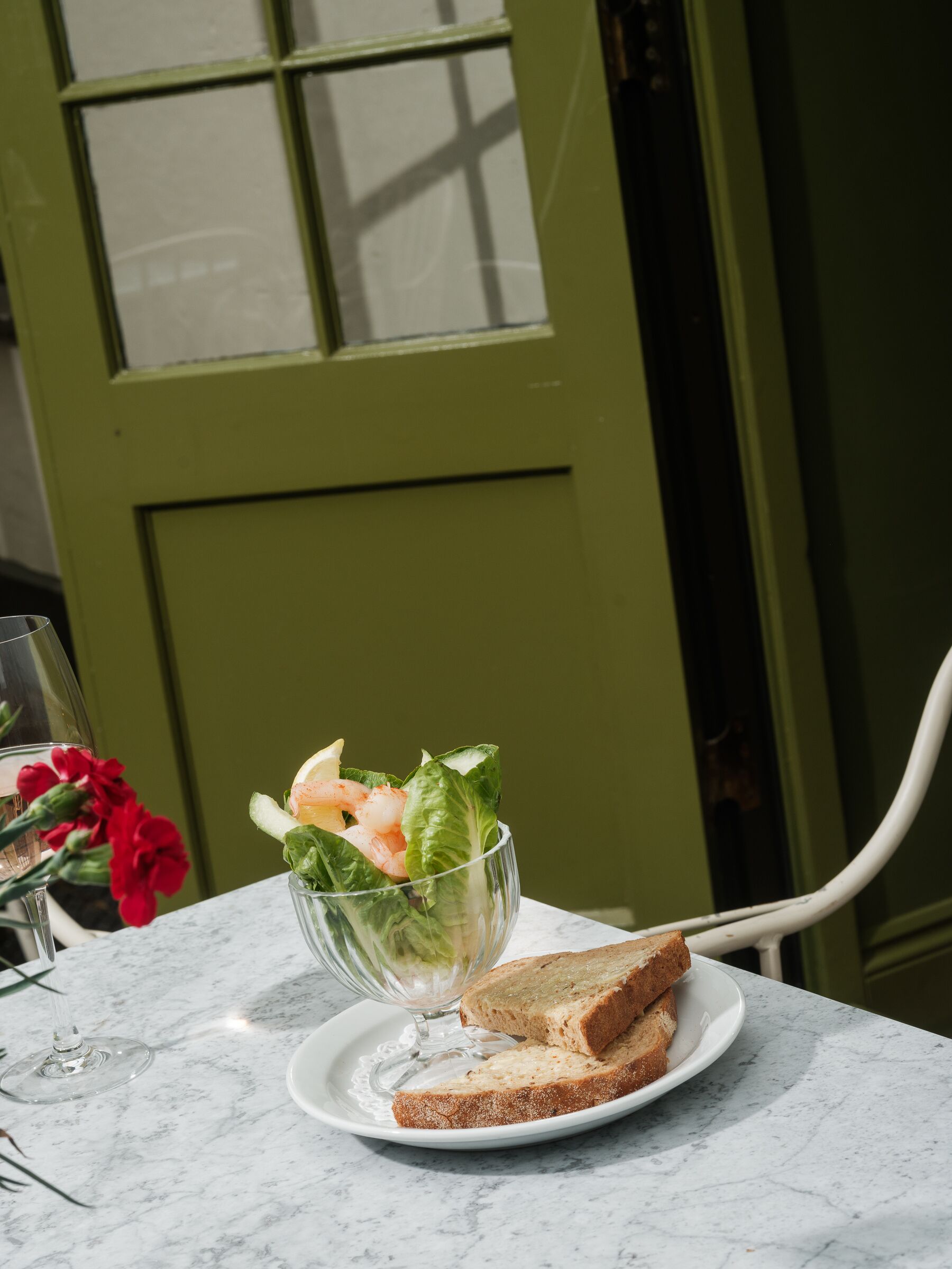 A clear glass bowl with a salad on a white plate, a wine glass, and a slice of bread on a marble table.