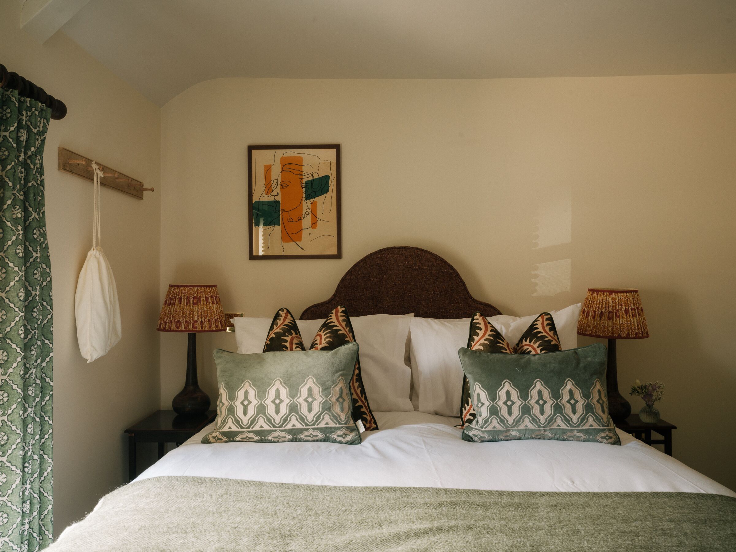 A neatly made bed with patterned pillows, a hanging white bag, two bedside lamps, and a framed abstract painting on the wall.