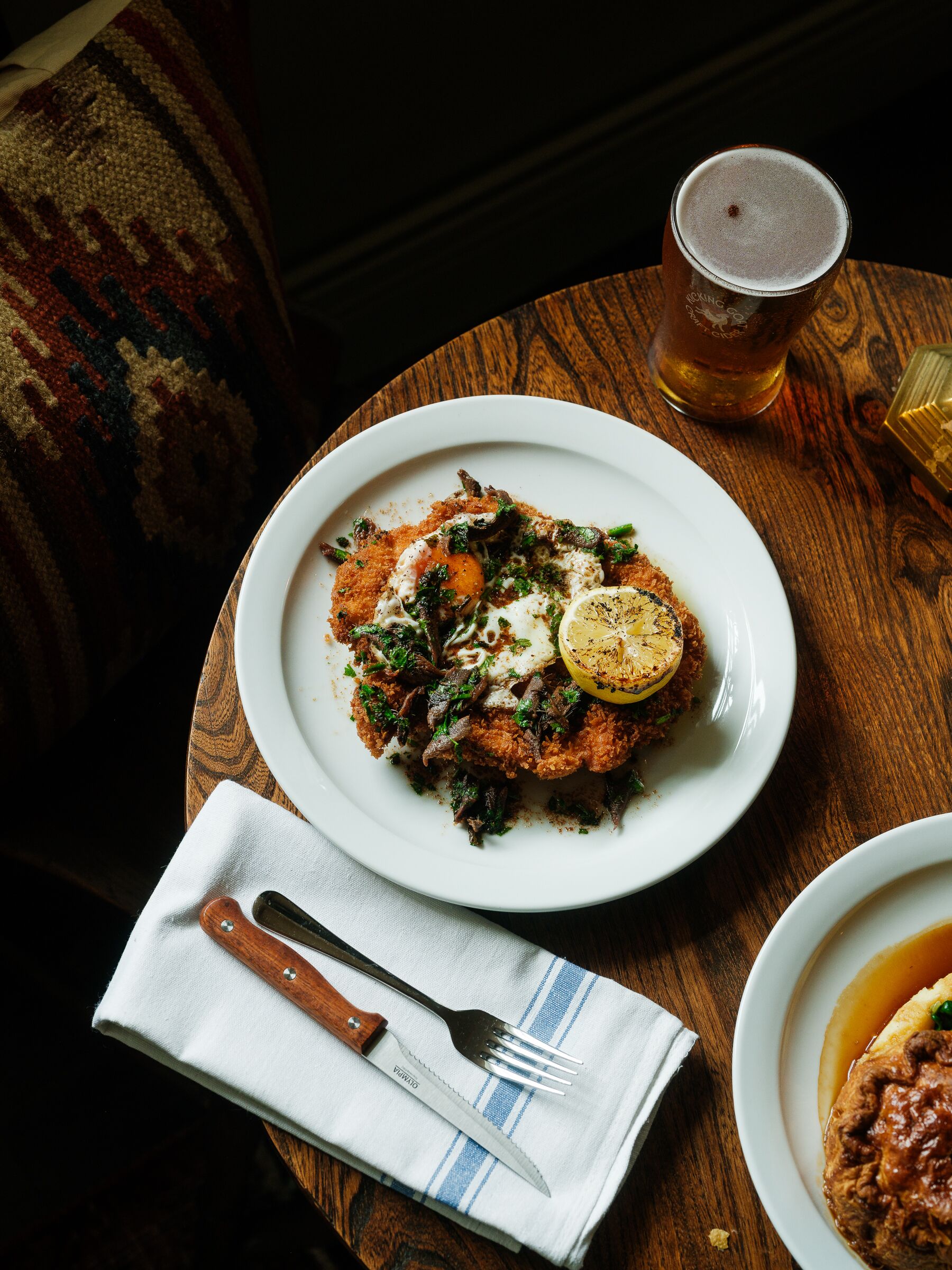 Un plato de comida con tenedor y cuchillo sobre una servilleta, una jarra de cerveza y un sofá estampado al fondo.