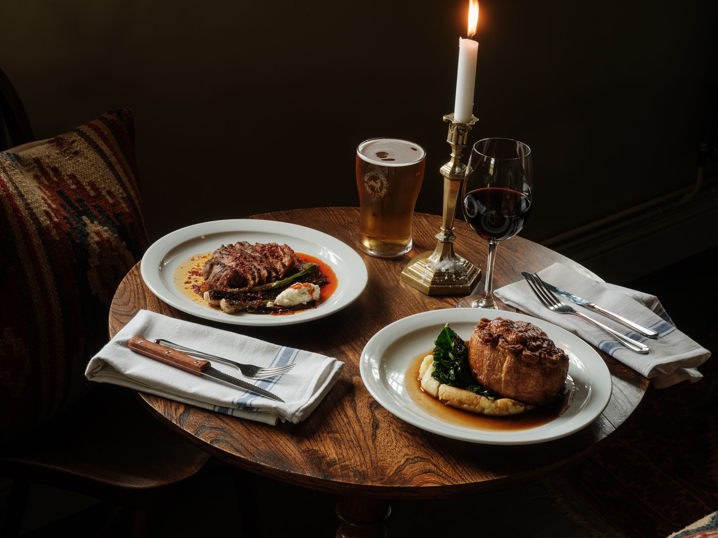 A dining table with two plates of food, a candle, a glass of beer, and a glass of red wine.