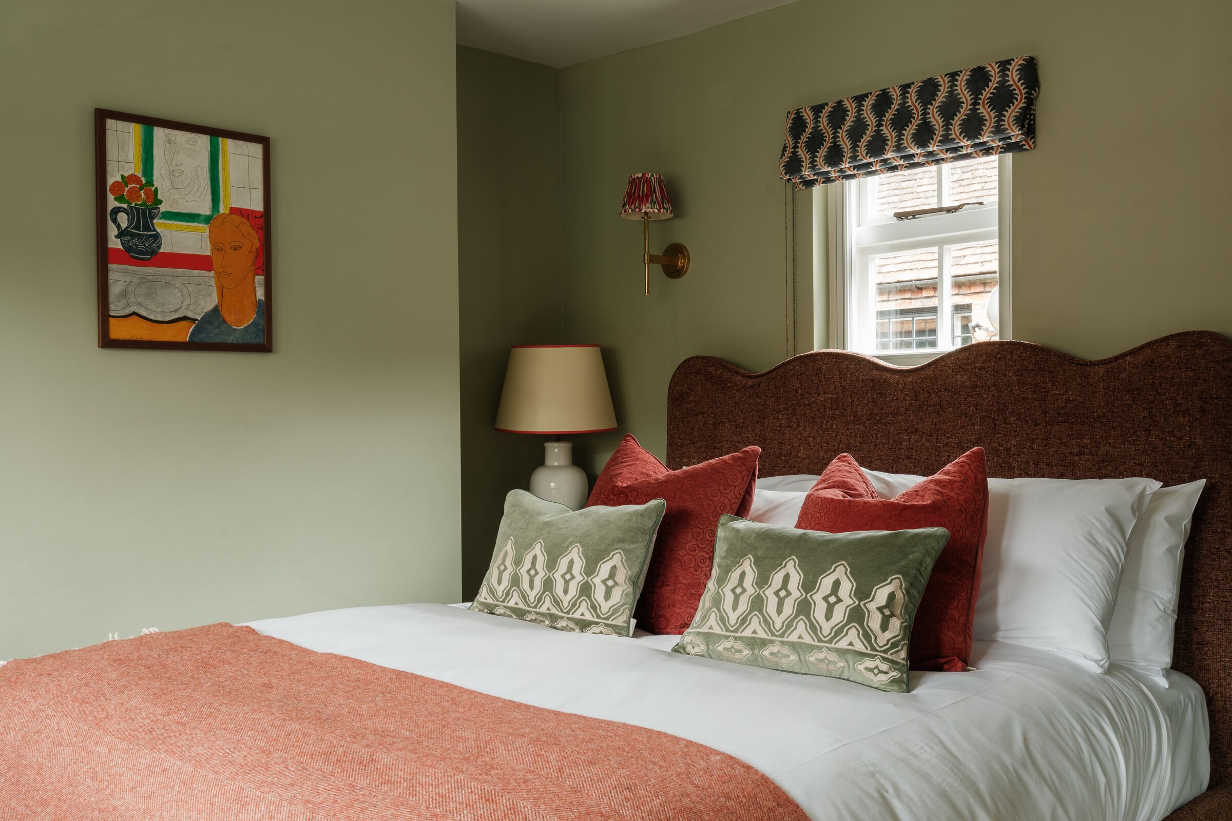 A bedroom with a bed, pillows, a lamp, a wall painting, and a window with a valance.