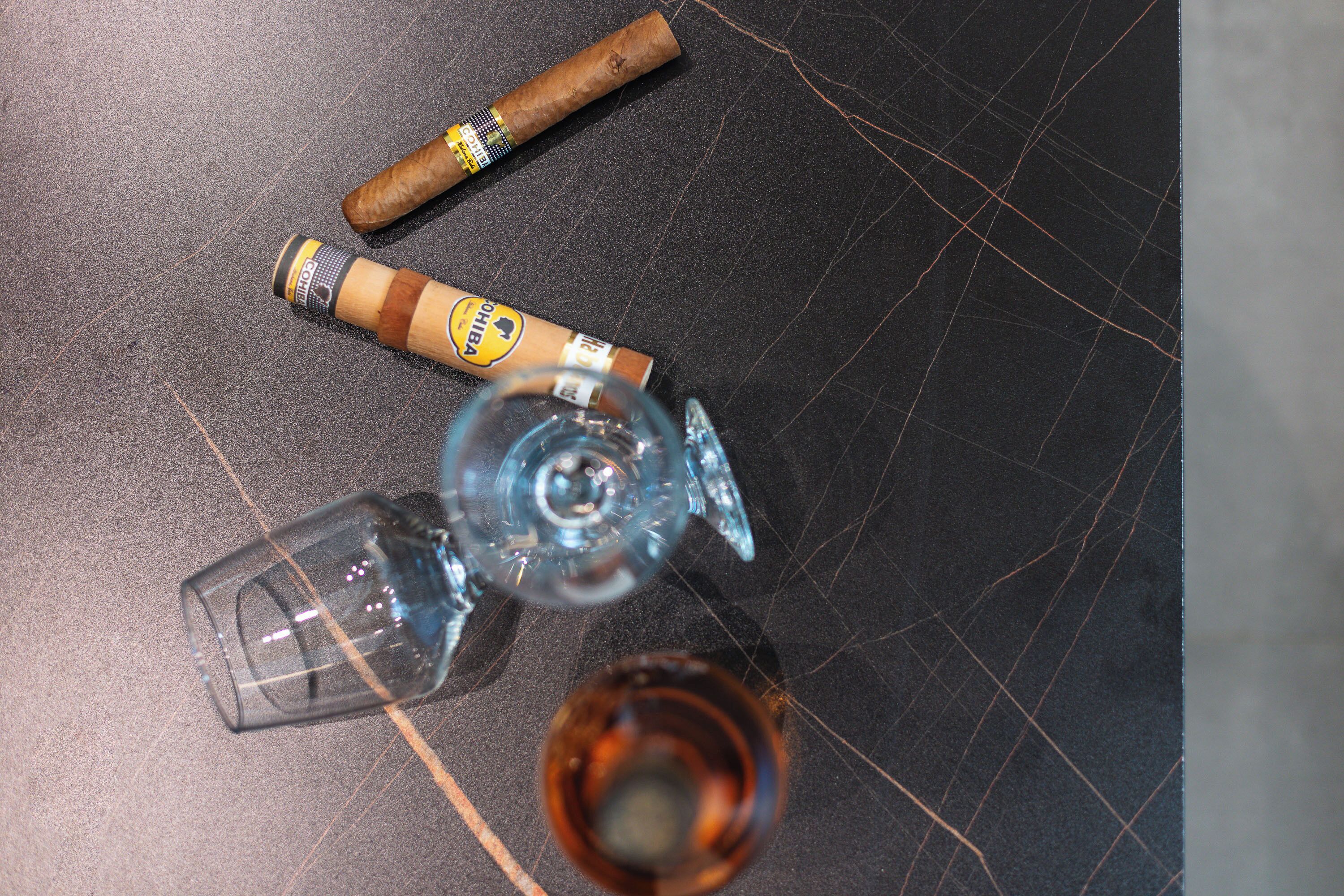 A glass bottle, a glass, and a cigar on a dark surface.