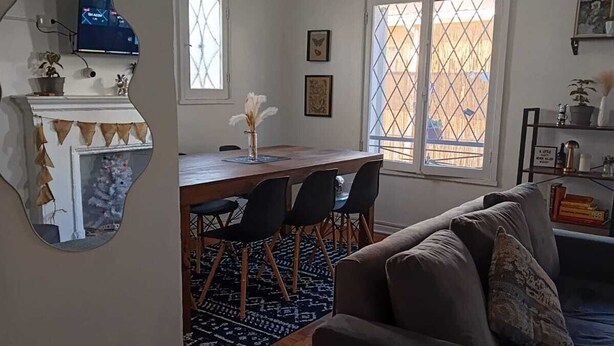 A living room with a grey sofa, a dining area with a wooden table and chairs, a fireplace, and a large mirror.
