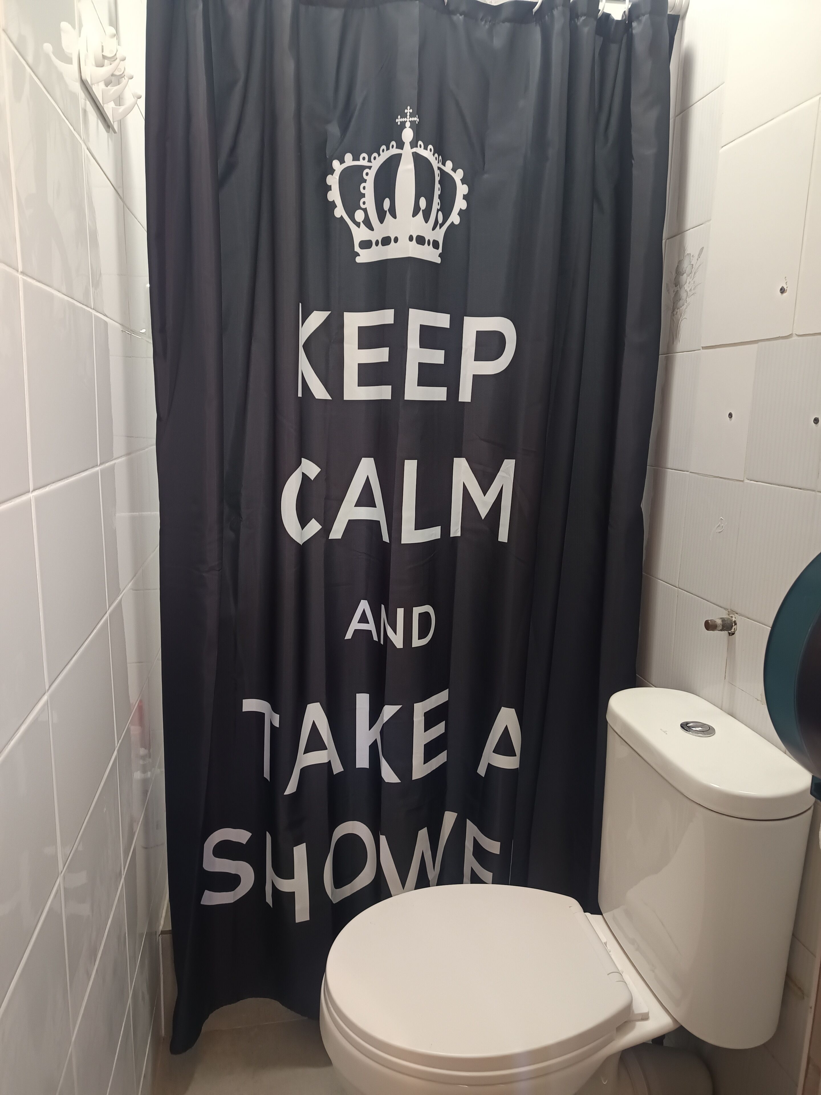 A bathroom with a black shower curtain featuring white text and a crown graphic, a white toilet, and white tiled walls.