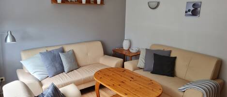 Una sala de estar con un sofá color beige, una mesa de centro de madera, una araña, una estantería de madera con objetos decorativos y una pequeña mesa redonda con un florero.