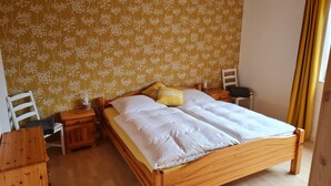 Un lit en bois avec une literie blanche et jaune, une table de chevet en bois, une chaise et un mur orné d’un motif floral.