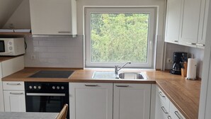 Eine kleine Küche mit weißen Schränken, einer eingebauten Mikrowelle, einem Fenster mit Blick auf Grünflächen und einer Kaffeemaschine.