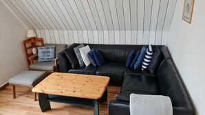 Ein modernes Wohnzimmer mit einem schwarzen Ledersofa, einem Holztisch und einem grauen Sessel. Ein Schild an der Wand zeigt 'The Lobster House'.