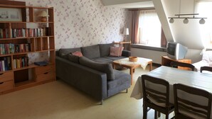 A living room with a sofa, a bookshelf, a dining table with chairs, a television, and a window with curtains. - Apartment 'Fewo Mirja' with Shared Terrace, Shared Garden and Wi-Fi (Pellworm)