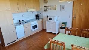 Een keuken met een spoelbak, fornuis, magnetron en kasten. Er staat een eettafel met stoelen en een groene tafelkleed.
