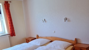 Een houten bed met twee kussens, een nachtkastje, een raam met rood gordijn en een radiator.