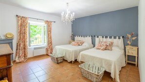 A bedroom with two beds, a chandelier, a window with curtains, a small bedside table, and a basket. - Gite le Bananier – 6 guests (Jonquières)