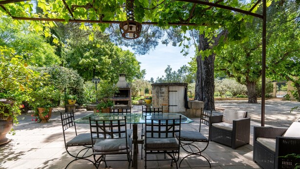 Un patio al aire libre con una mesa de vidrio, sillas de mimbre y una zona de barbacoa de piedra.