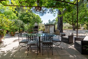 An outdoor patio with a glass table, wicker chairs, and a stone barbecue area. - Gite le Grenadier – 6 Guests (Jonquières)