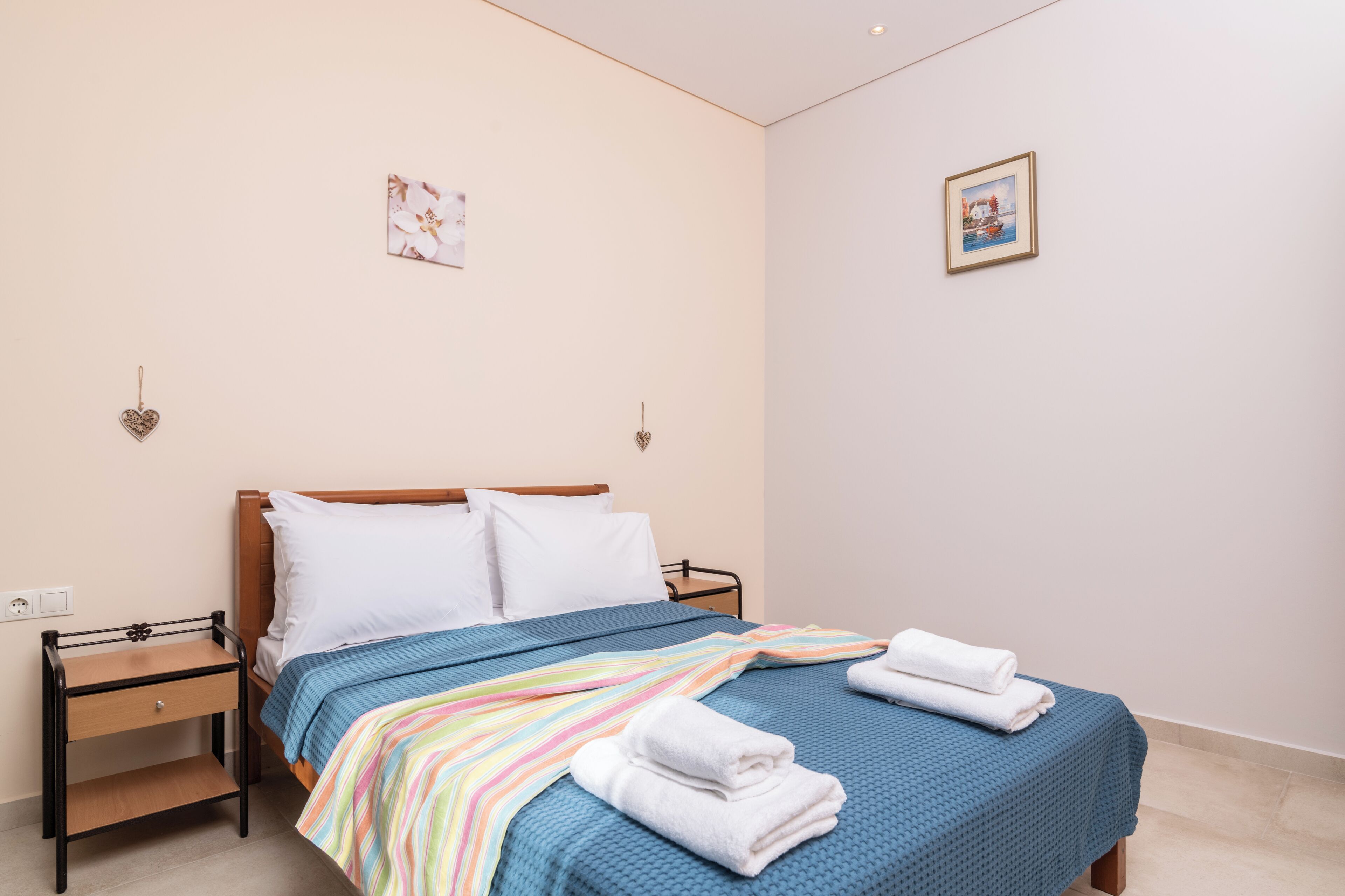 A neatly made bed with a blue bedspread and white pillows, a bedside table with a drawer, and two wall-mounted heart-shaped decorations.