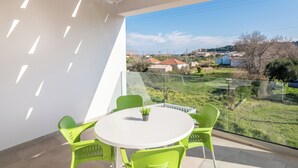 Ein Balkon mit einem weißen Tisch und grünen Stühlen, der auf eine grüne Fläche mit Häusern und einem Gewässer blickt.