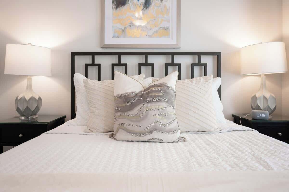 Una cama bien hecha con ropa de cama blanca y almohadones decorativos, flanqueada por dos mesitas de noche con lámparas y un reloj.