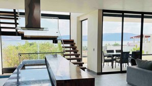 A modern kitchen with a central island, a staircase leading to a balcony, and a seating area with a view of the outdoors. - Casa Cuba - Spectacular Lake and Mountain View (Jocotepec)