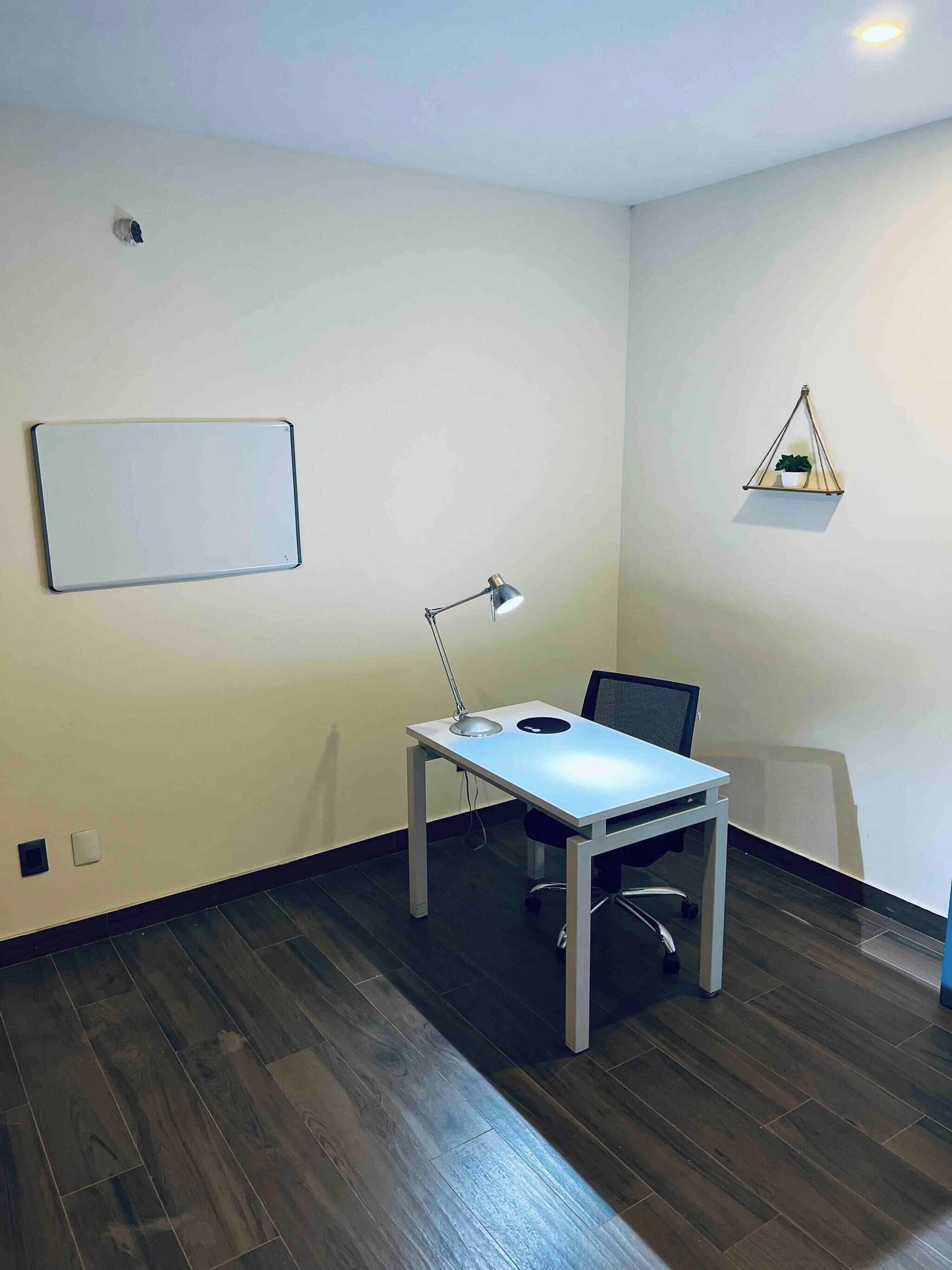 A small office space with a desk, chair, whiteboard, desk lamp, and a hanging plant.