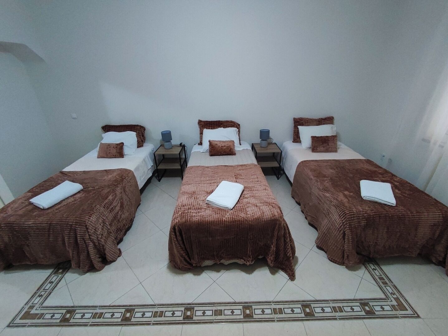 A room with three single beds, each with a pillow and a folded towel. There are bedside tables with lamps.