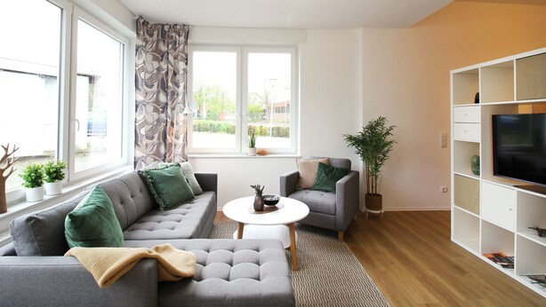 Una sala de estar moderna con un sofá y un sillón grises, una mesa de centro blanca y un televisor sobre un mueble blanco. Hay plantas en macetas y una ventana con cortinas.