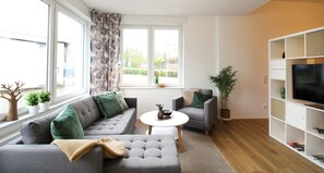 Un salon moderne avec un canapé gris, un fauteuil gris, une table basse blanche et un téléviseur posé sur un meuble blanc. On y trouve également des plantes en pot et une fenêtre avec des rideaux.