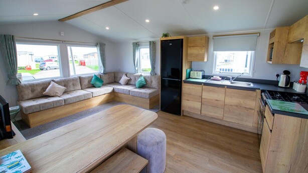 A compact RV interior with a sofa, dining table, kitchen area, and large windows. - MP416 Parkdean Resorts Camber Sands (Camber)
