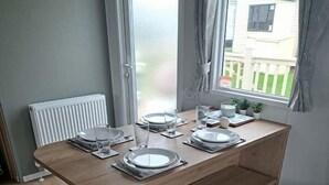 A modern dining area with a wooden table set for four, two grey ottomans, a white radiator, and a window with curtains. - MP416 Parkdean Resorts Camber Sands (Camber)