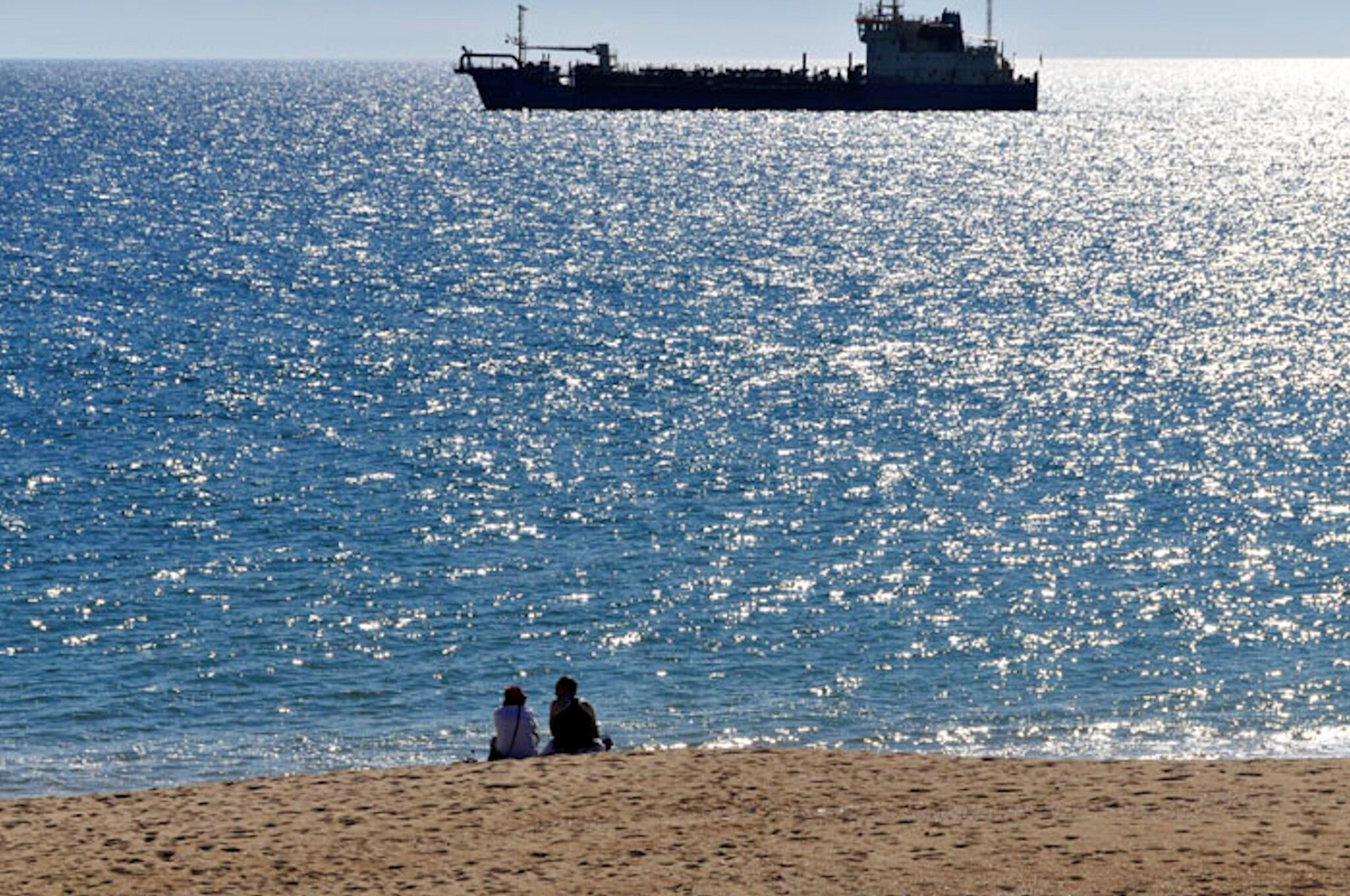 A beach with two people sitting on the sand, a large ship in the water, and a clear sky.