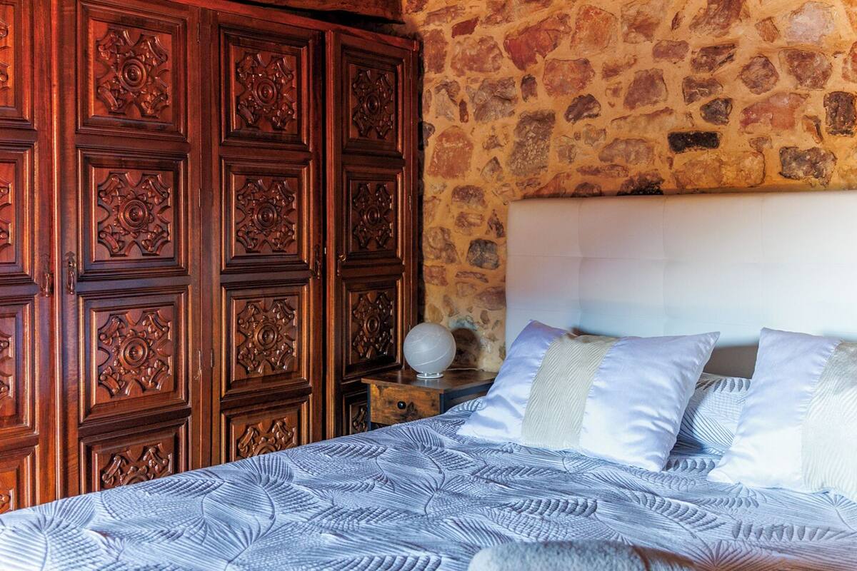 A bedroom with a stone wall, wooden wardrobe, and a bed with patterned bedding.