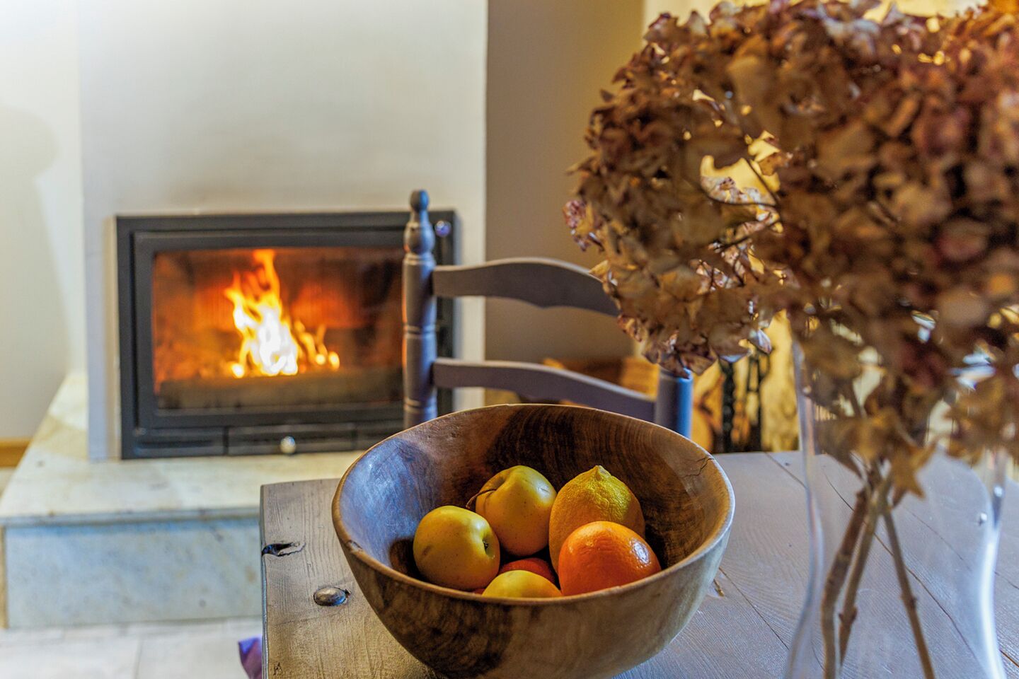 Un cuenco de madera con frutas sobre una mesa, una silla y una chimenea encendida.