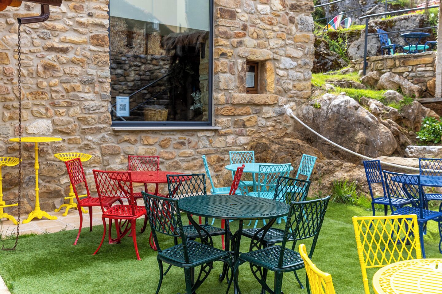 Un edificio de piedra con una ventana, mesas y sillas coloridas en una terraza, y un área con césped.
