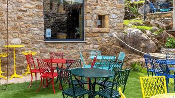 Un edificio de piedra con una ventana, mesas y sillas coloridas en una terraza, y un área con césped.