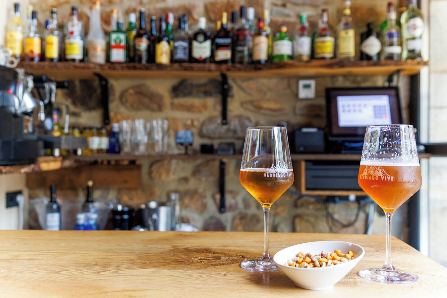 Two glasses of beer on a wooden table with a bowl of nuts beside them, and a bar with various bottles in the background.