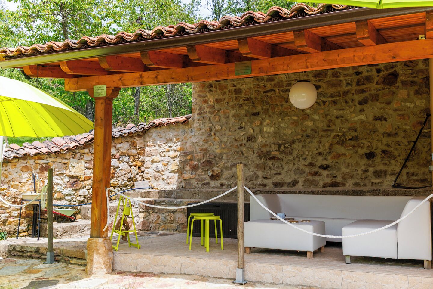 A stone-walled outdoor seating area with a wooden pergola, a white sofa, and a yellow umbrella.