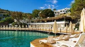 A poolside area with lounge chairs and umbrellas, a wooden deck, and buildings in the background. - Başka Resort Bodrum - All inclusive (Bodrum)