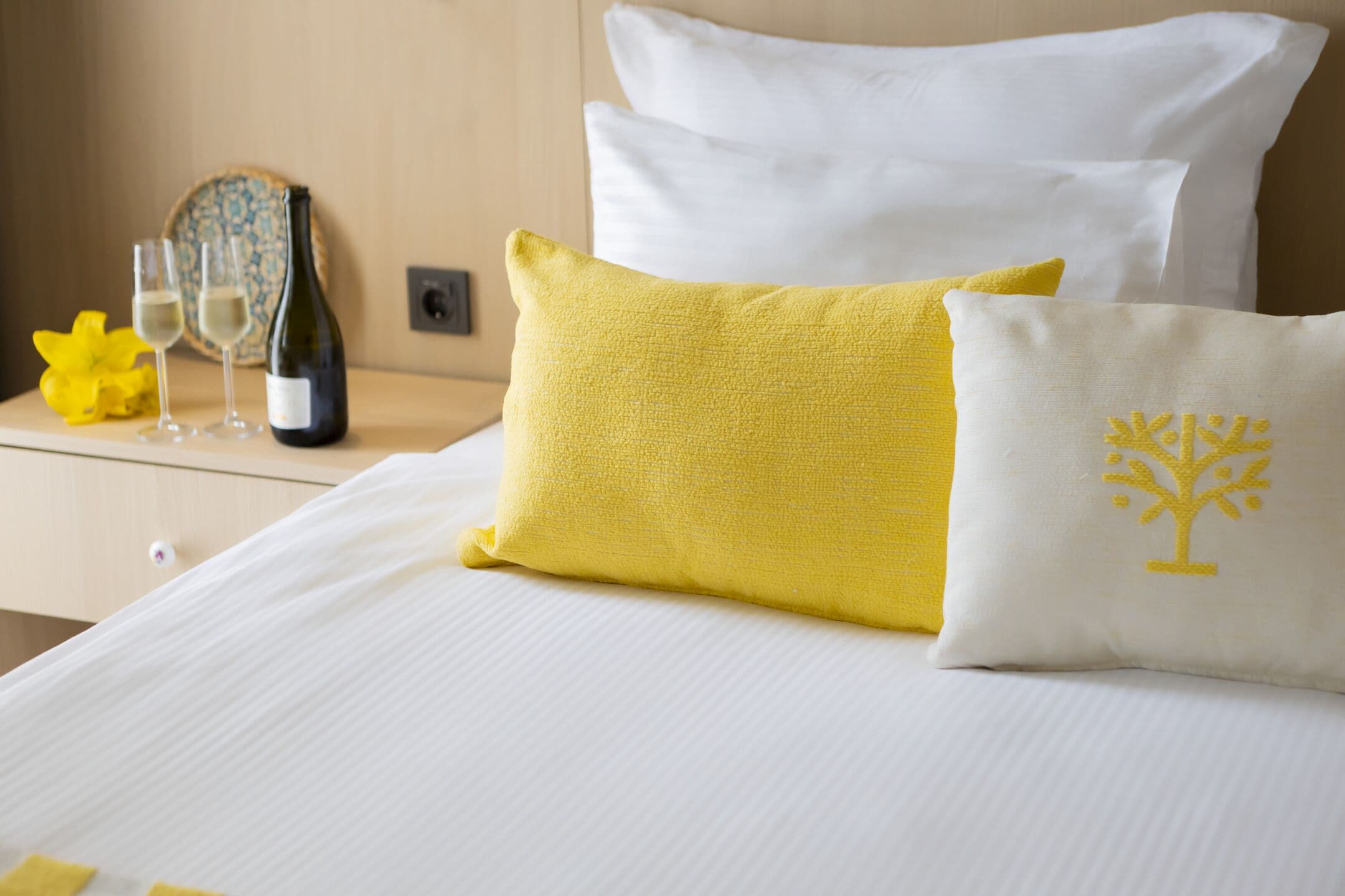 Habitación de hotel con una cama bien hecha, una mesita de noche con una botella de vino y copas, y una flor amarilla decorativa.