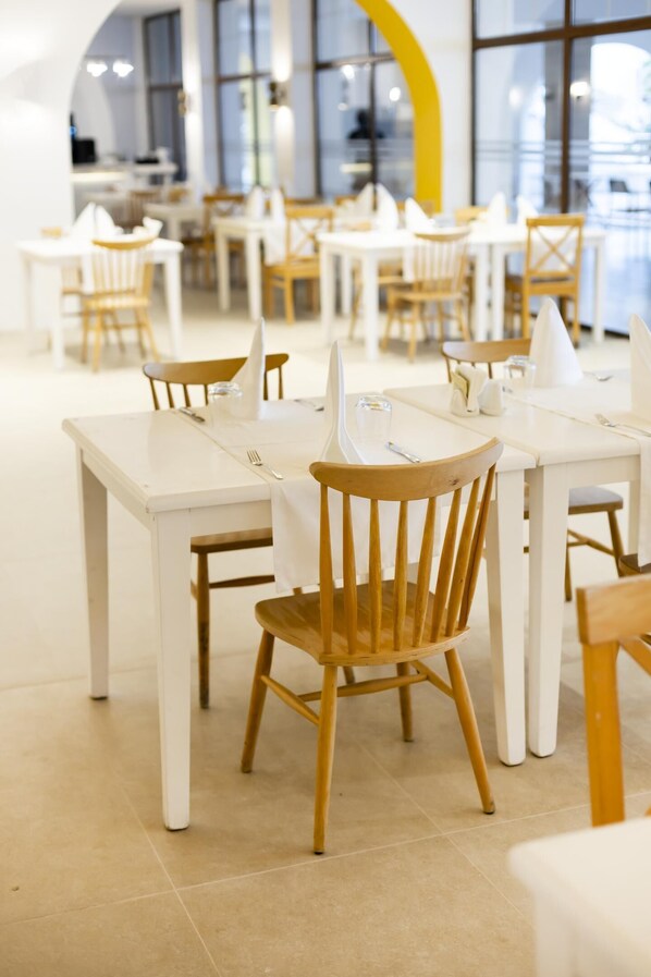 Un comedor moderno con mesas blancas y sillas de madera, ventanas amplias y una pared de acento amarilla.