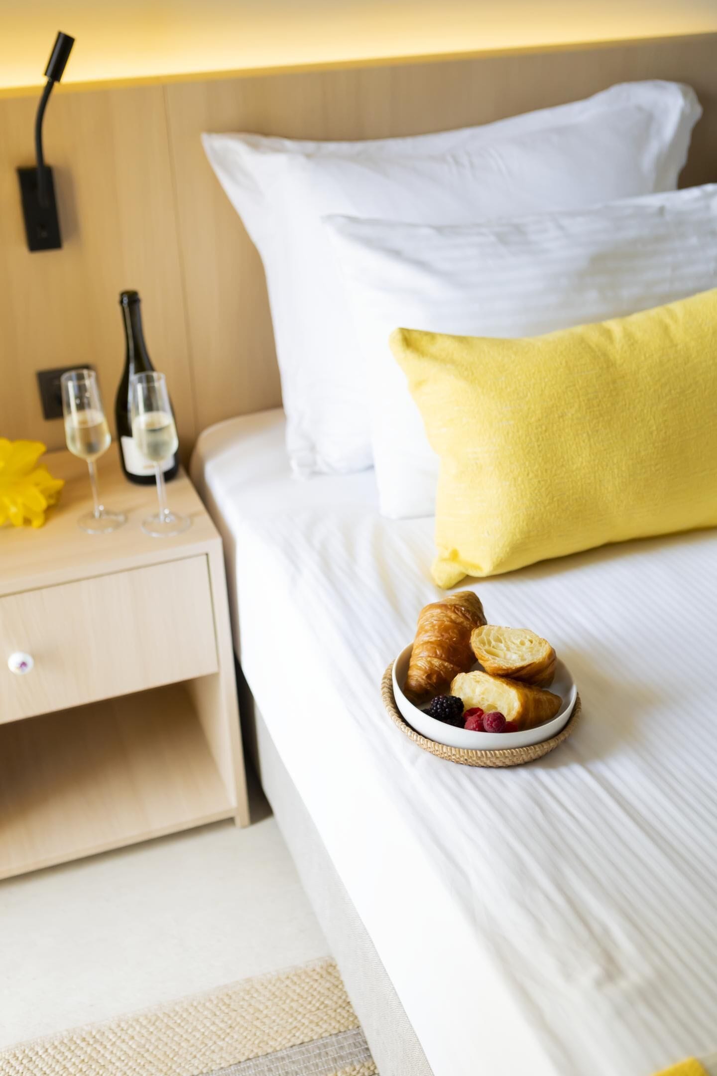 A hotel room with a bed, a bedside table with two glasses of wine, a bottle, and a basket of pastries.
