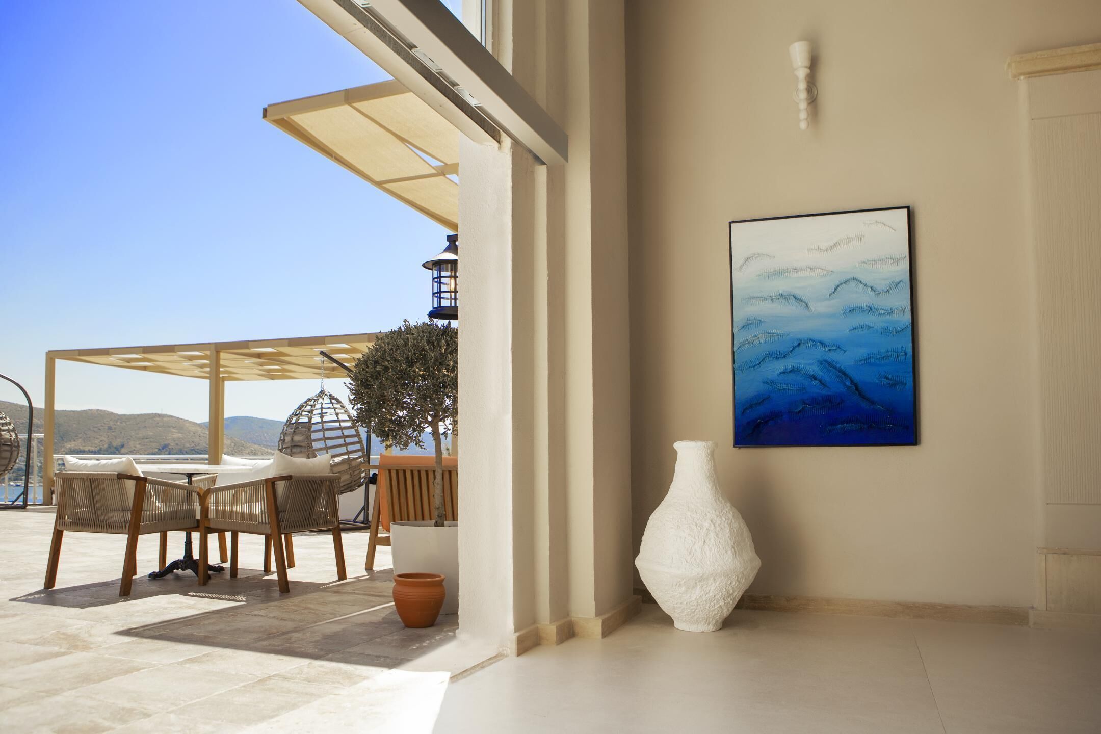 A sunlit room with a view of the sea, a table with chairs, a white vase, and a painting on the wall.