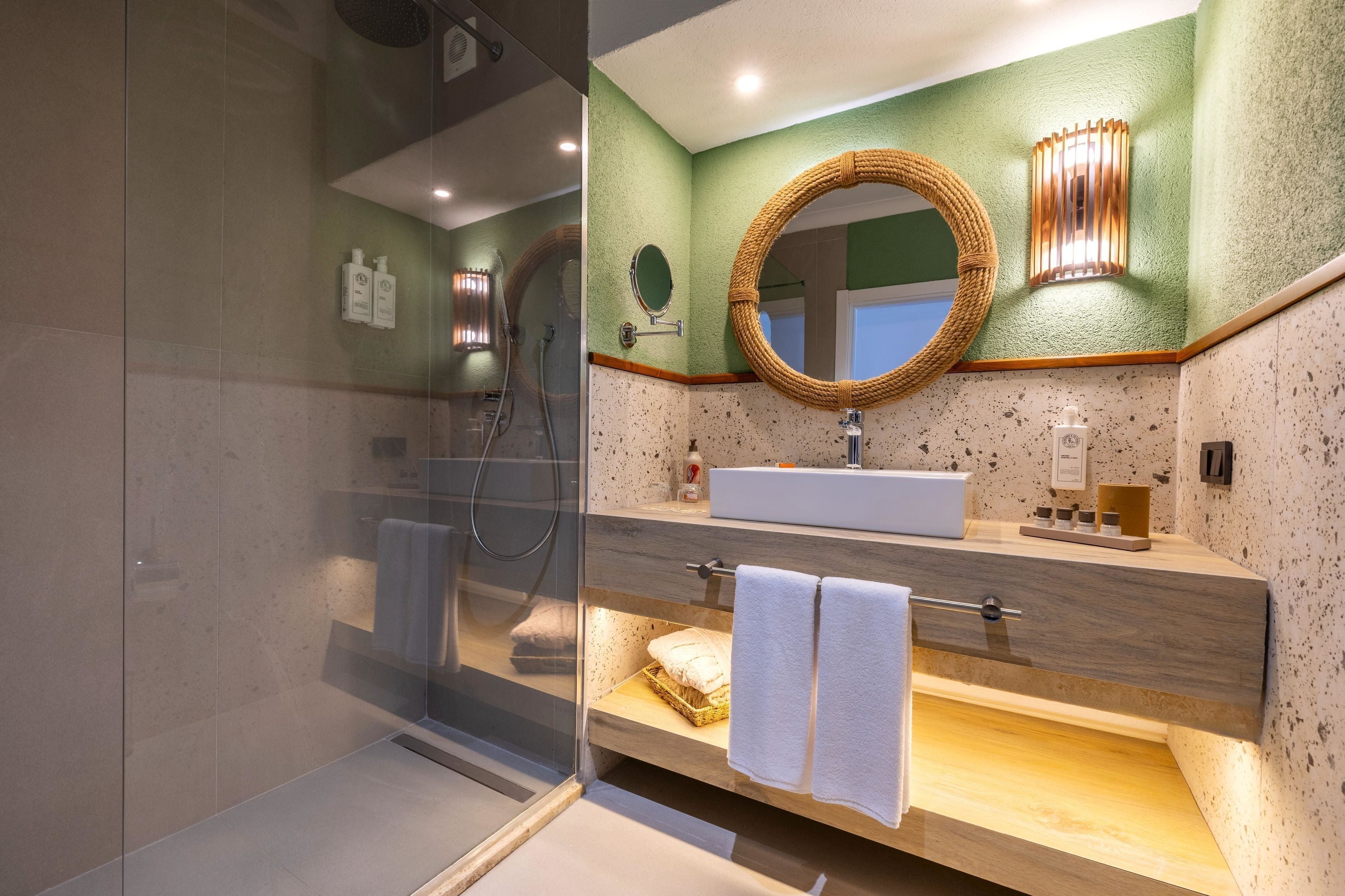 Un baño moderno con cabina de ducha de vidrio, tocador de madera con lavamanos y espejo con marco de fibras naturales.