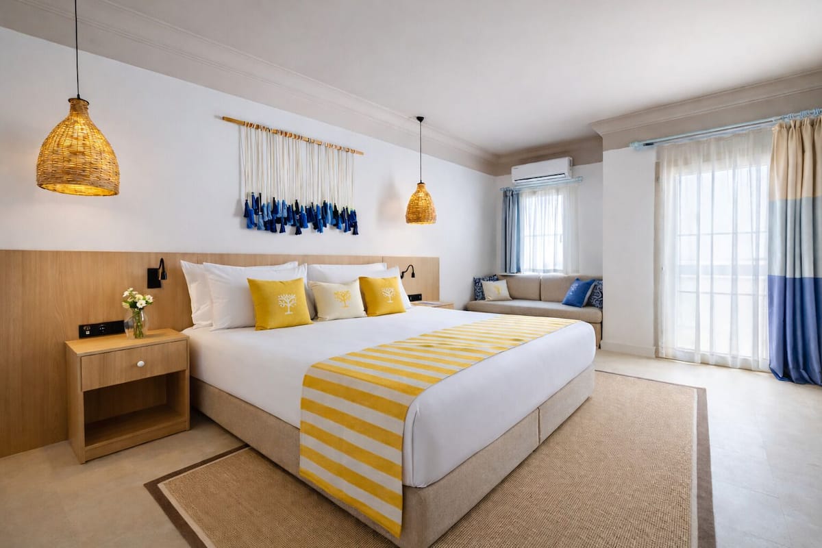 Habitación de hotel con una cama grande, mesitas de noche, un sofá, una lámpara colgante y una ventana con cortinas.