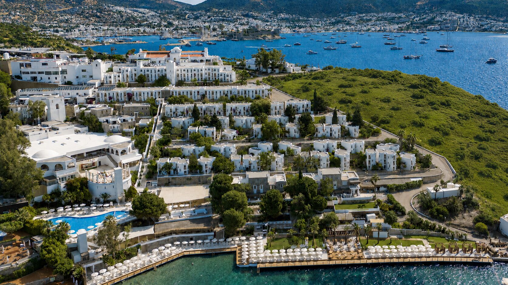 Une vue aérienne d’une ville côtière avec des bâtiments blancs, un espace piscine central et des bateaux sur l’eau.