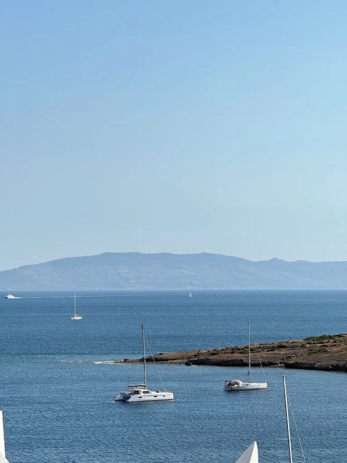 Escena costera con varios veleros anclados en aguas tranquilas, cielo despejado y montañas a lo lejos.