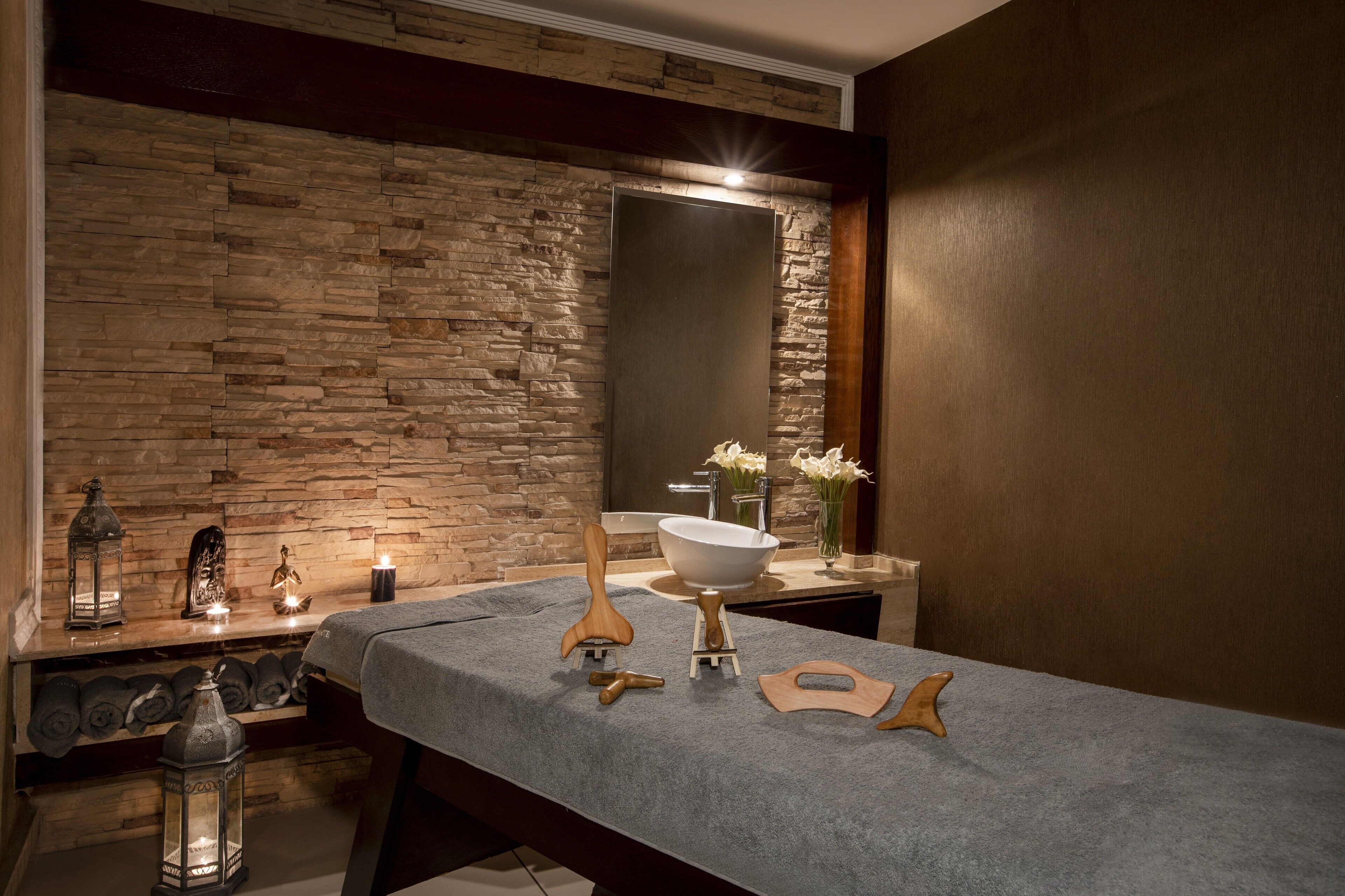 A modern bathroom with a stone wall, a large mirror, a white sink, and a wooden massage table.