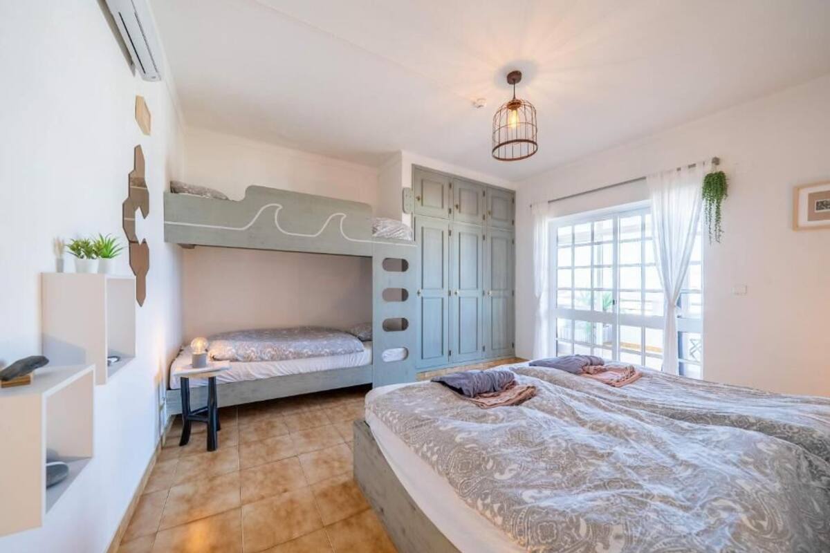 Un dormitorio con una cama litera, una cama grande, un armario, una mesita de noche y una planta colgante.
