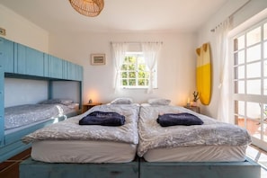 A room with two beds, a window with curtains, a surfboard on the wall, and a woven light fixture. - SURFinn Algarve (Aljezur)